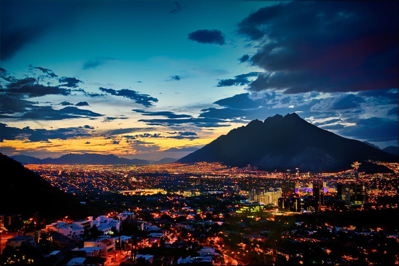 Panorámica nocturna de la ciudad de Monterrey, México, con montañas al fondo y un cielo azul lleno de nubes y tonos anaranjados.