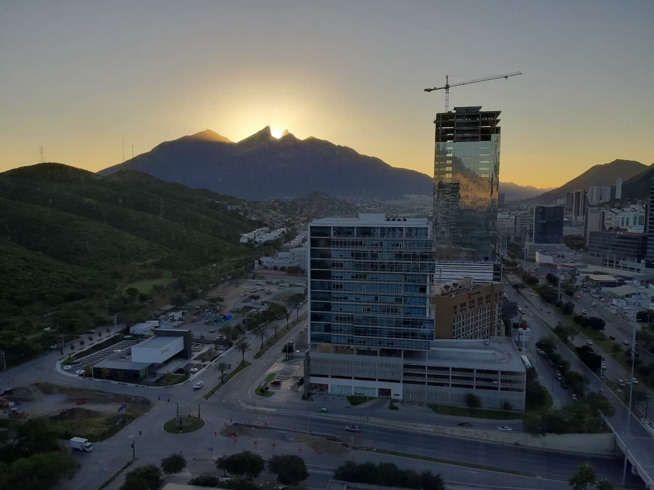 Vista panorámica de Monterrey al atardecer, con montañas al fondo, edificios modernos y una grúa de construcción.