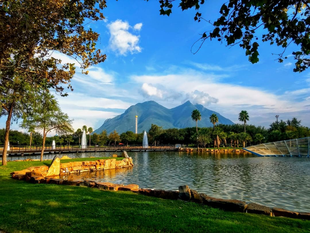 Parque con lago, montañas al fondo, palmeras y fuentes en el agua.