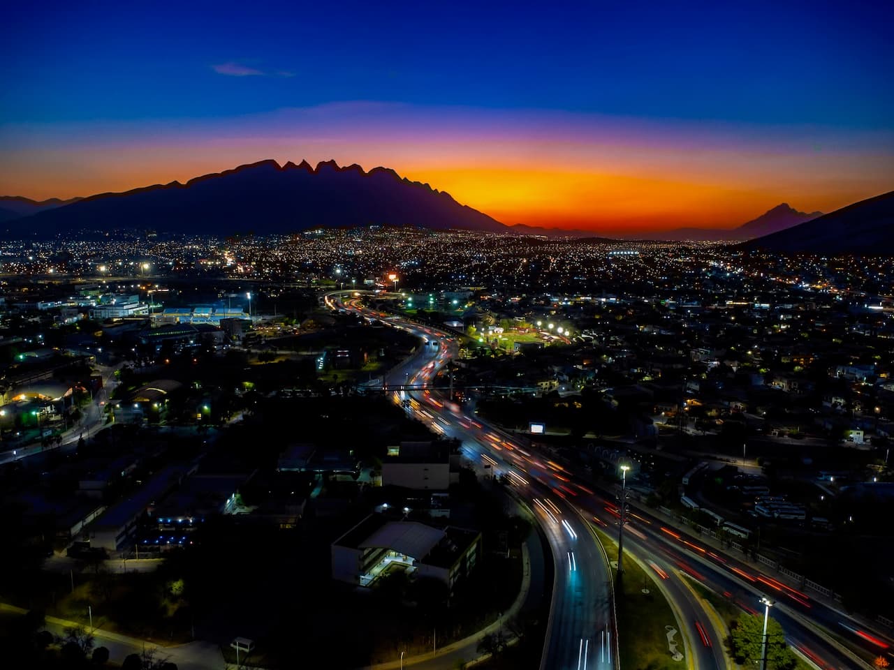 Vista nocturna de una ciudad con montañas al fondo, donde se aprecian luces urbanas y tráfico en movimiento en las carreteras.