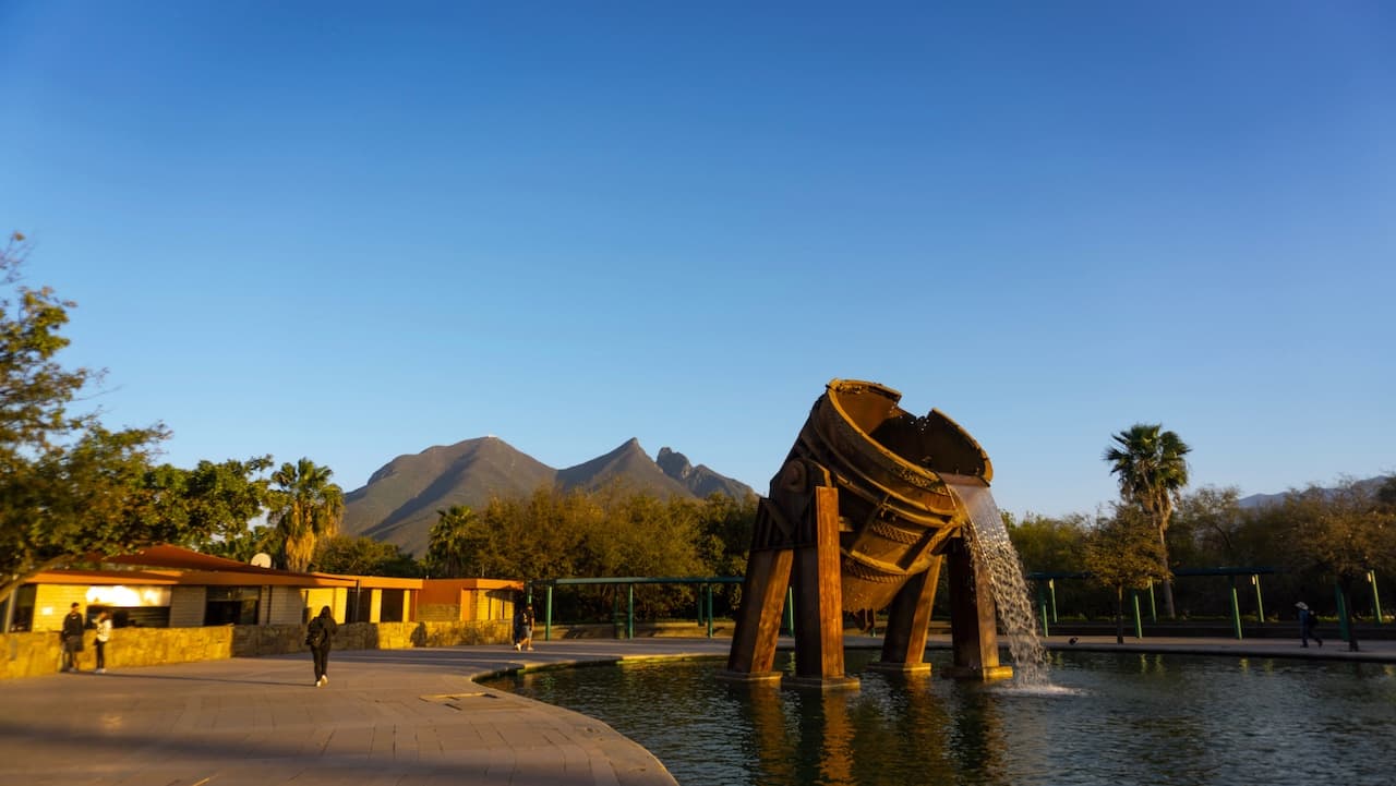 Escultura de una gran cubeta vertiendo agua en un estanque, con montañas al fondo y árboles alrededor.