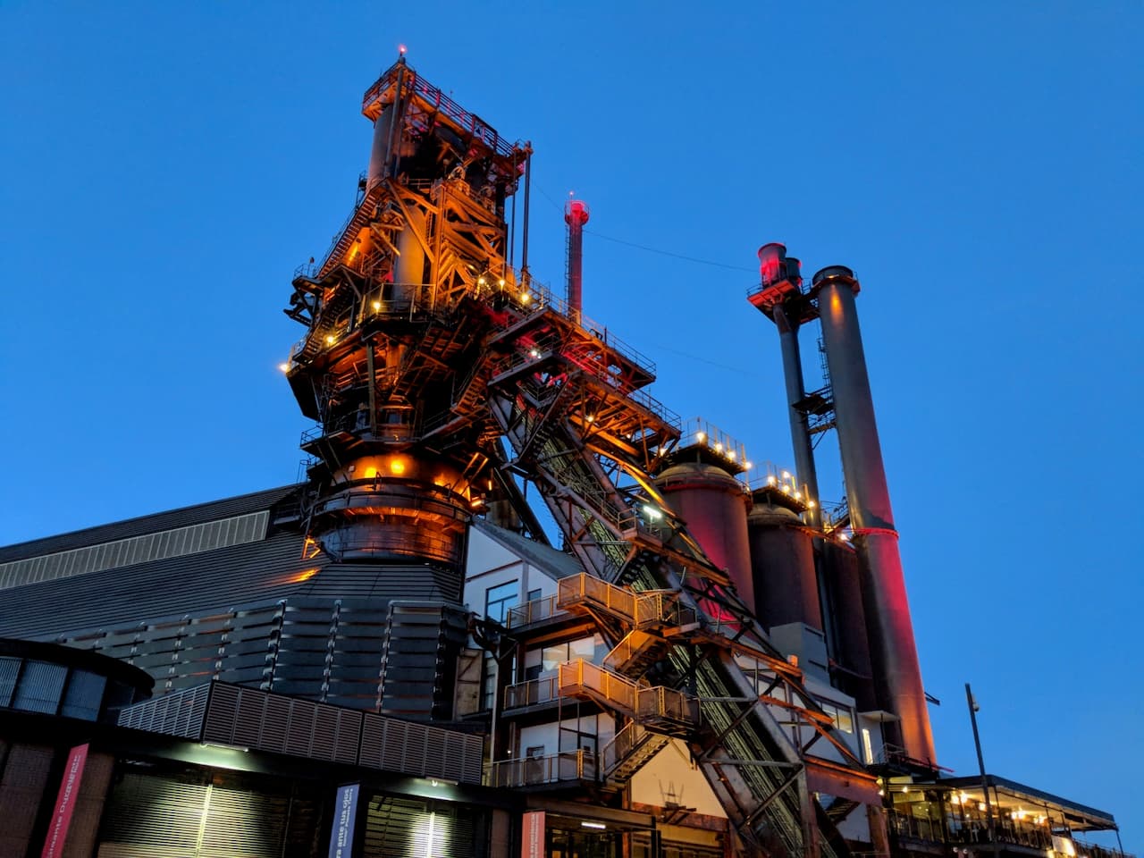 Planta industrial con estructuras metálicas iluminadas, en un fondo de cielo azul al atardecer.