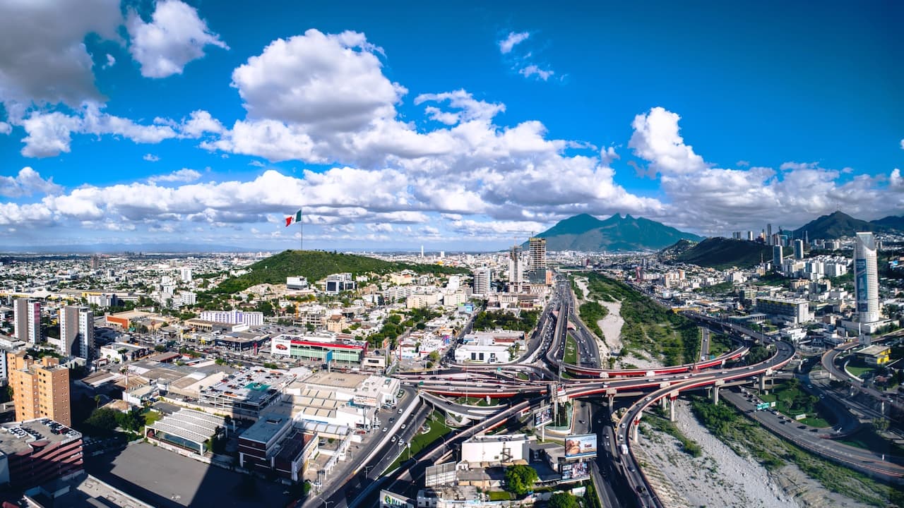 Vista panorámica de una ciudad con edificios, montañas y un cielo con nubes. Se observa una carretera con tráfico y una gran bandera mexicana ondeando.